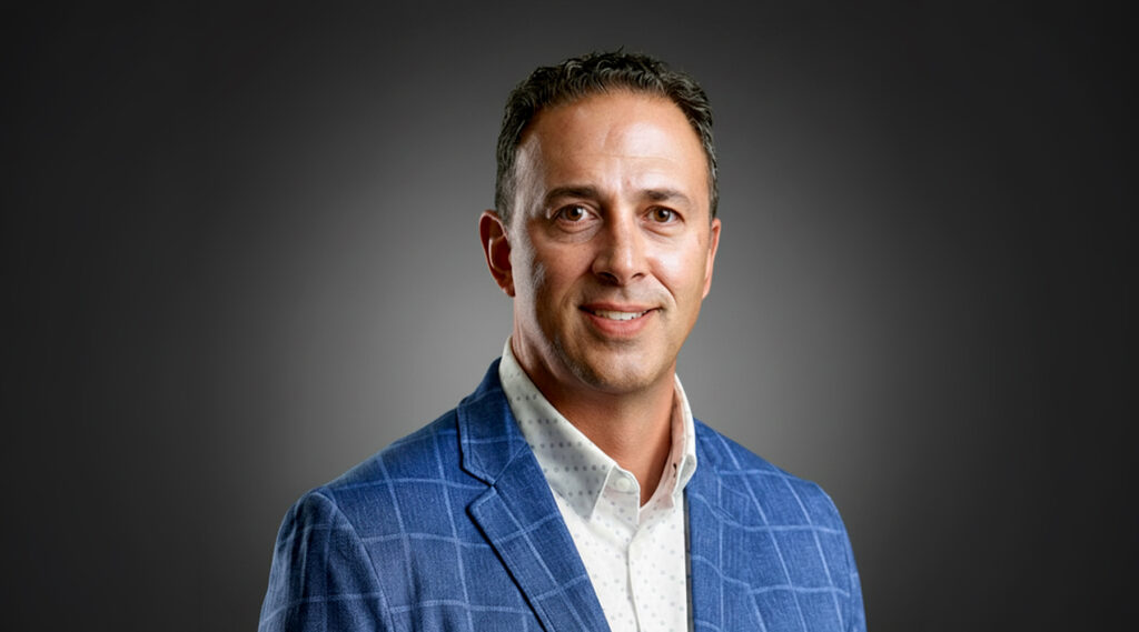 Smiling man with short dark hair in a blue checkered blazer and white shirt against a plain dark background.