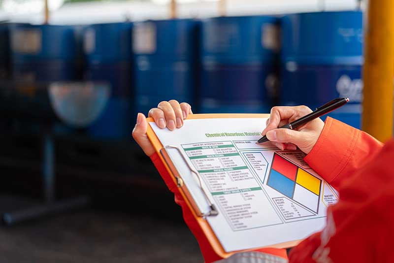 Worker in orange safety gear filling out a chemical hazard form on clipboard near blue chemical barrels in storage area.