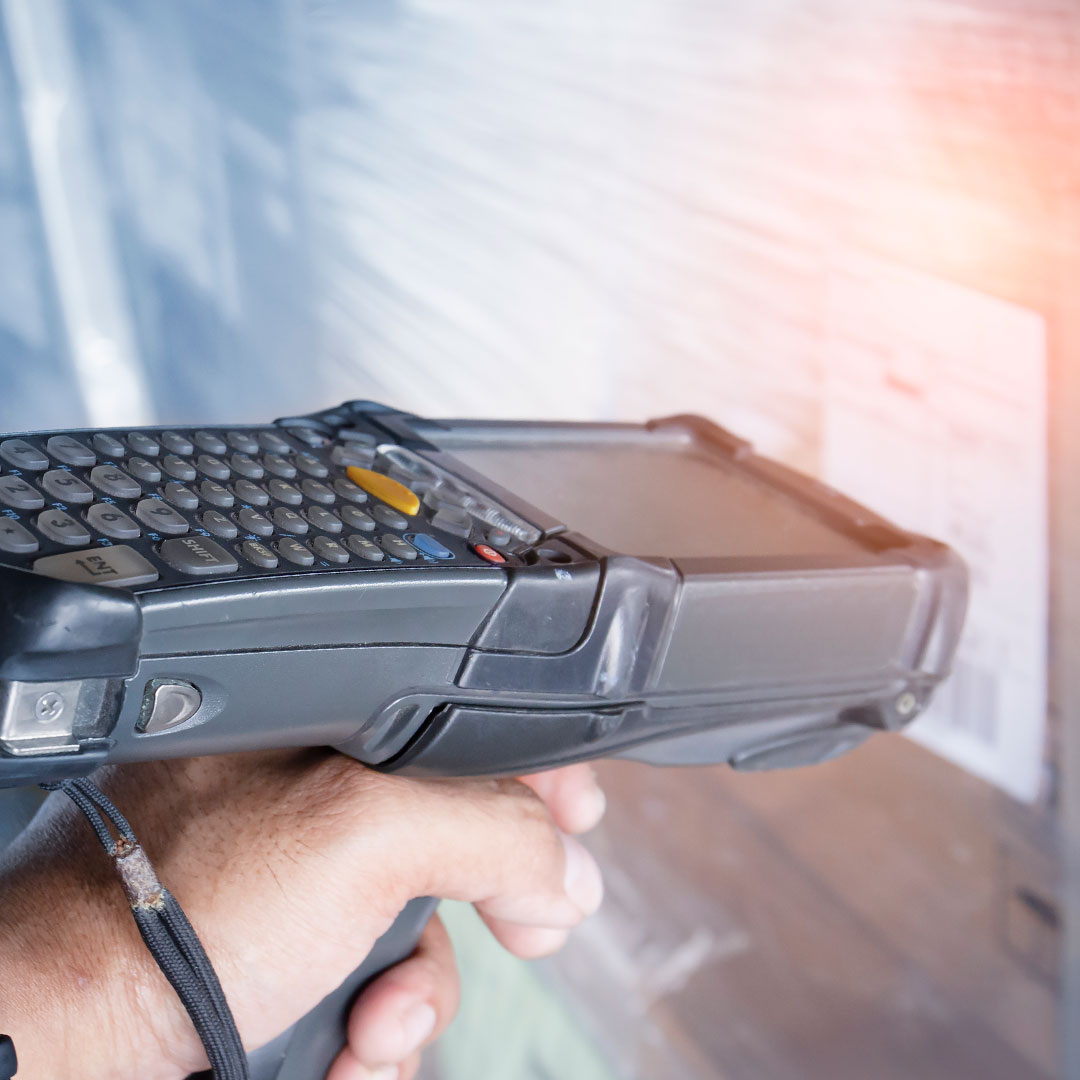 A hand holds a barcode scanner with a keypad and screen, scanning a label on a wrapped pallet or package in a warehouse setting. Light reflects off the wrapped surface.