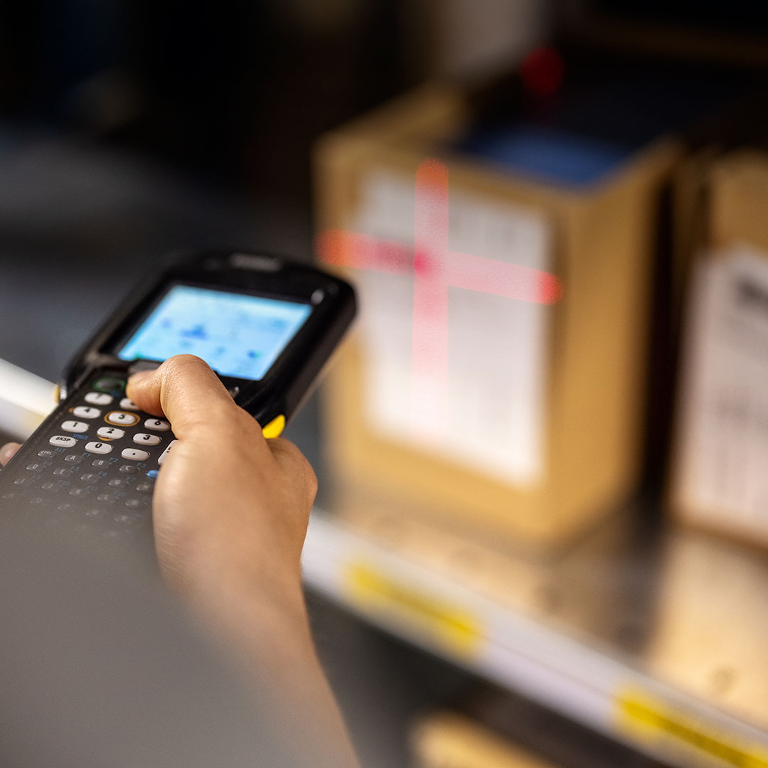 Person scans a cardboard box label with a handheld barcode scanner and red laser cross in an organized warehouse setting.