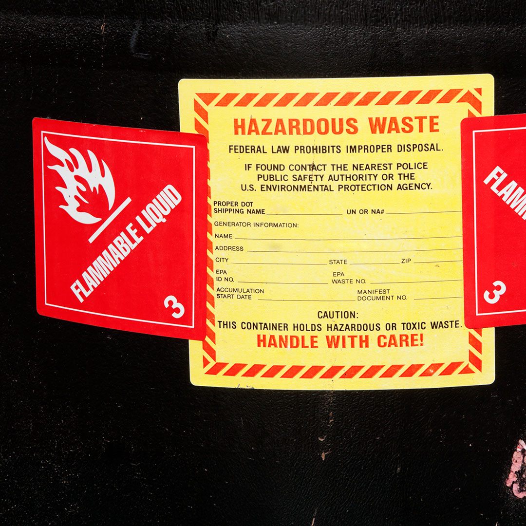 Yellow hazardous waste label with red border, handle with care text, flanked by red flammable liquid signs on dark surface.