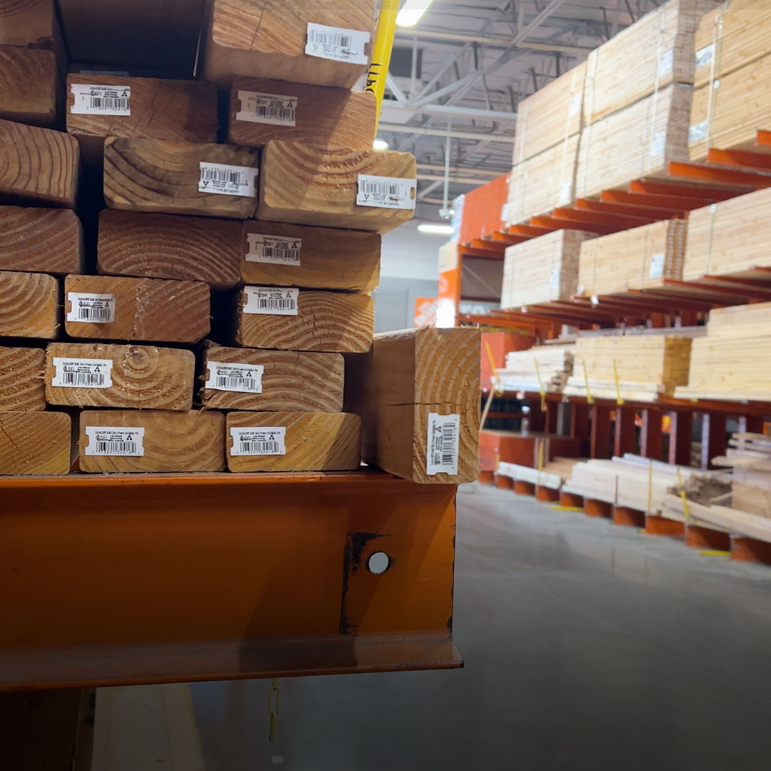 Stacked lumber with barcodes on orange shelves in a hardware store aisle, additional wood planks visible in the background.
