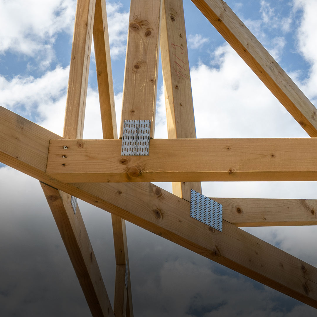 Wooden roof trusses with metal plates form a building framework under construction beneath a blue sky with scattered clouds.