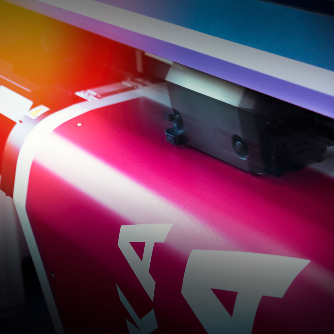 Large format printer producing pink and white graphics on paper, with motion blur and reflected colorful light in the background.