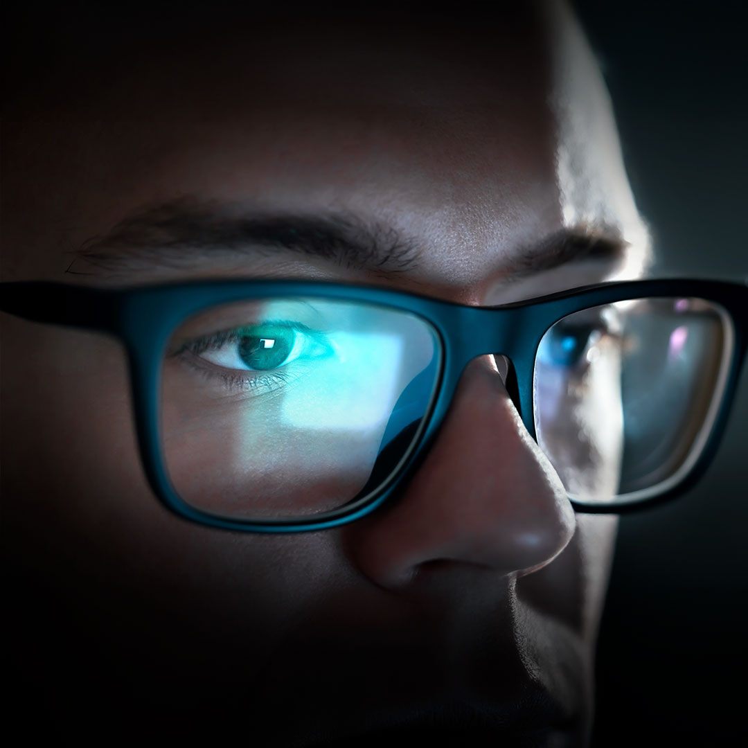 Person with glasses reflects screen’s blue light, focused eyes and serious expression visible in dim lighting.