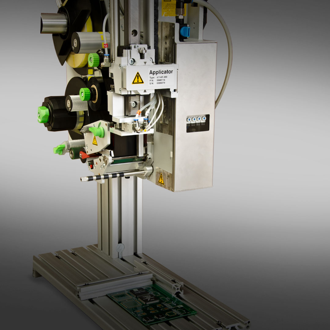 Industrial label applicator with rollers, control panel, and warning labels mounted above a green circuit board on a metal frame.