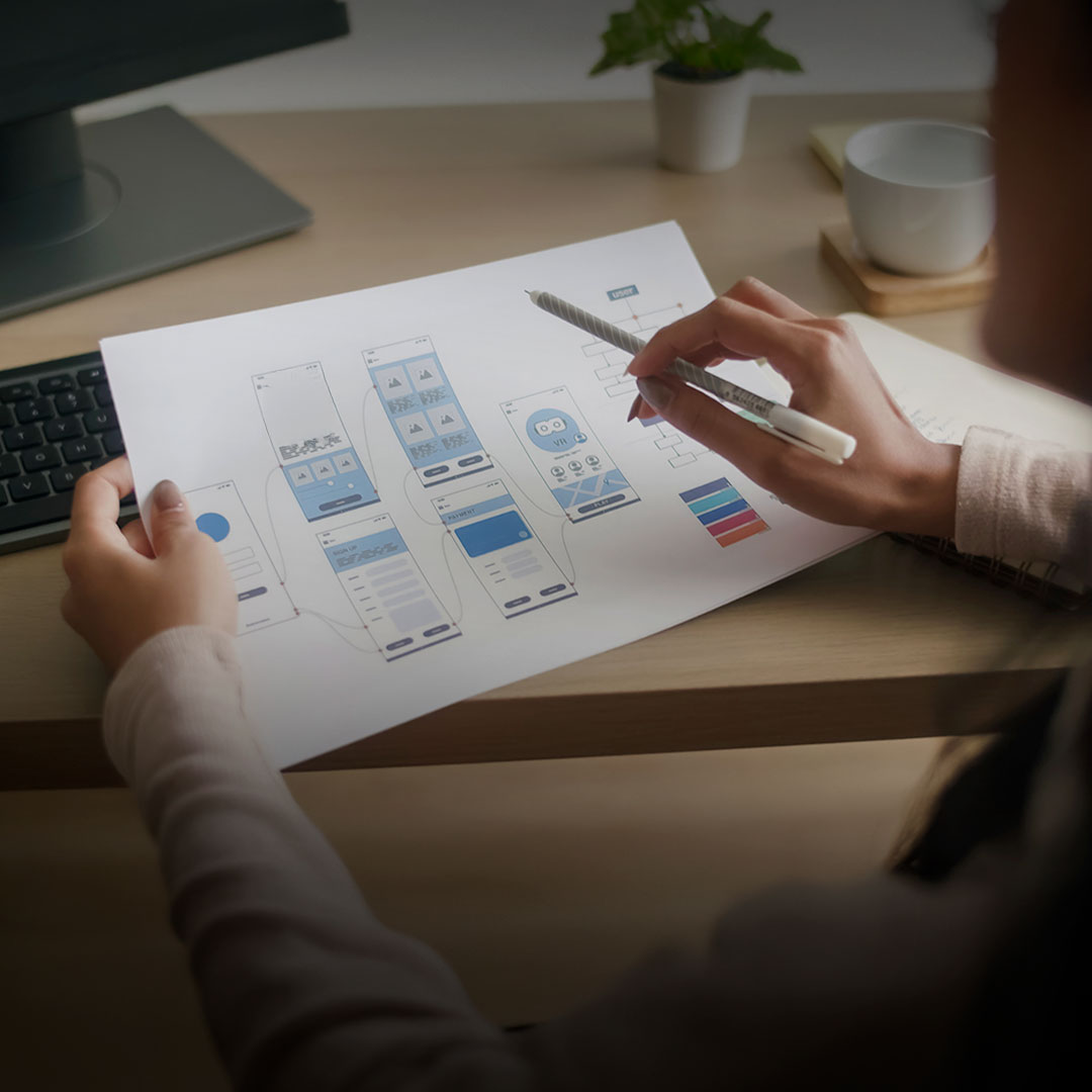 Person holding mobile app wireframes and design plans at a desk with keyboard, potted plant, and cup nearby.