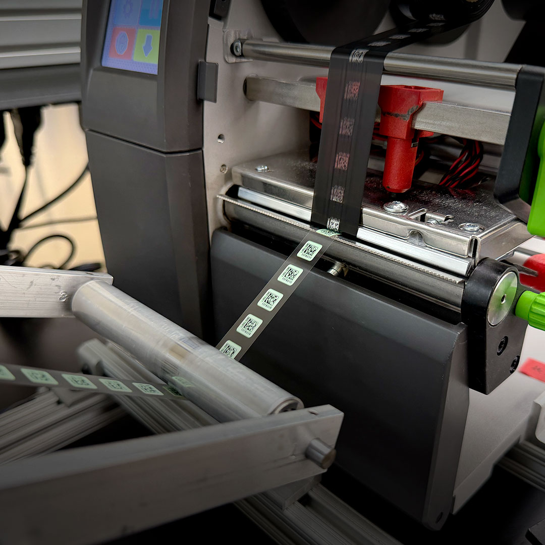 Close-up of a label printer producing small square barcode labels onto a roll in an industrial environment.