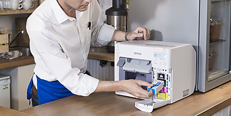 Person in white shirt and blue apron installs ink cartridge in Epson printer on wooden counter in kitchen or café setting.