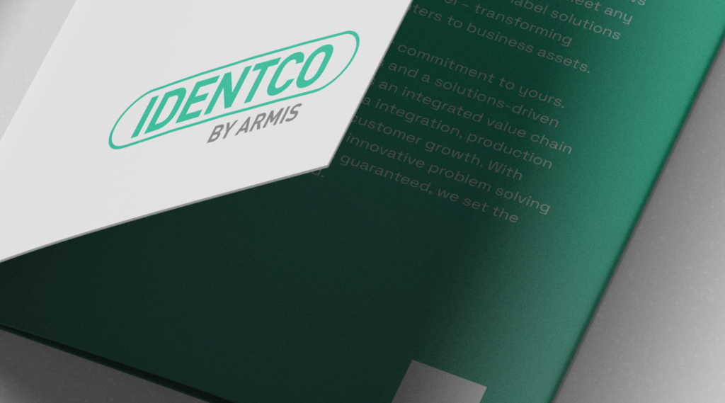 Brochure with IDENTCO BY ARMIS logo in green and gray on white, partly over green pages with text, placed on a gray surface.