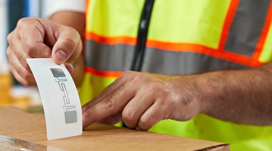 Worker in yellow safety vest with orange stripes presses RFID tag or label onto cardboard box using hands.