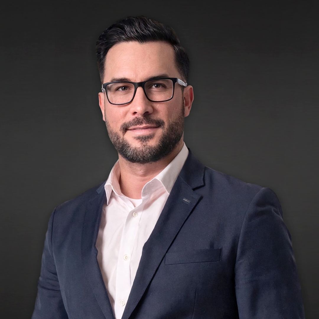 Confident man with short dark hair, beard, and glasses wearing navy blazer and white shirt against a plain dark background.