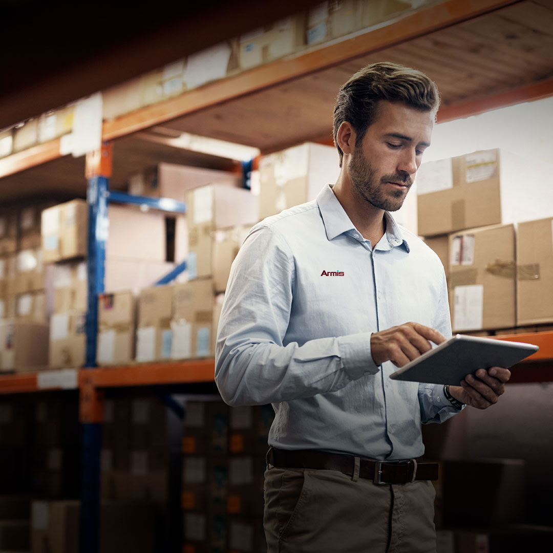 Man in a warehouse uses a tablet in front of shelves with cardboard boxes, wearing a light blue Armis logo shirt.