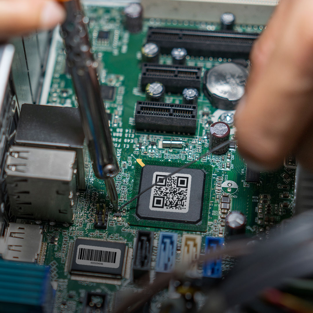 Hands use a tool on a computer motherboard with visible ports, chips, and a QR code on the central black chip.