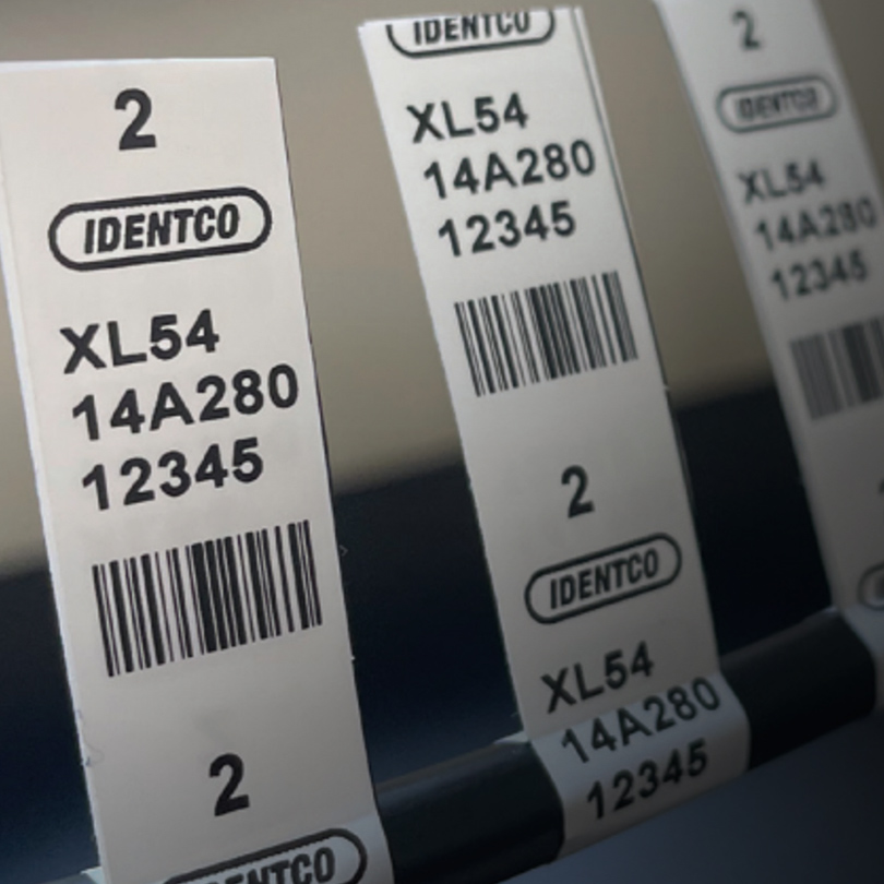 Close-up of white barcode labels IDENTCO, XL54, 14A280, 12345, each marked with number 2 and attached to a black organizer.