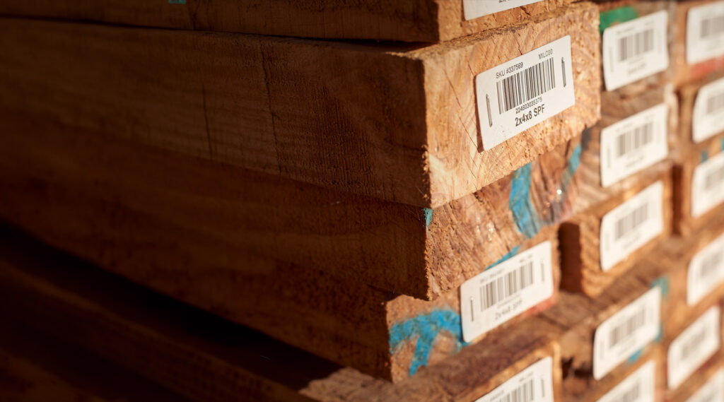 Stacked wooden planks with barcoded labels, size markings, and blue paint on edges under warm lighting.