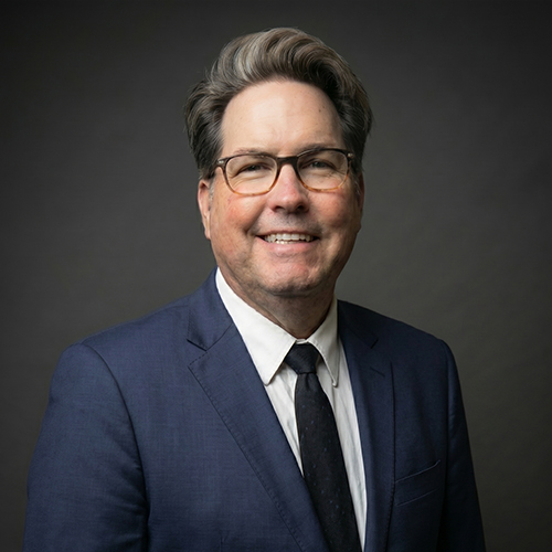 Frank, a smiling man in glasses, navy suit, white shirt, and black tie stands against a plain dark background.