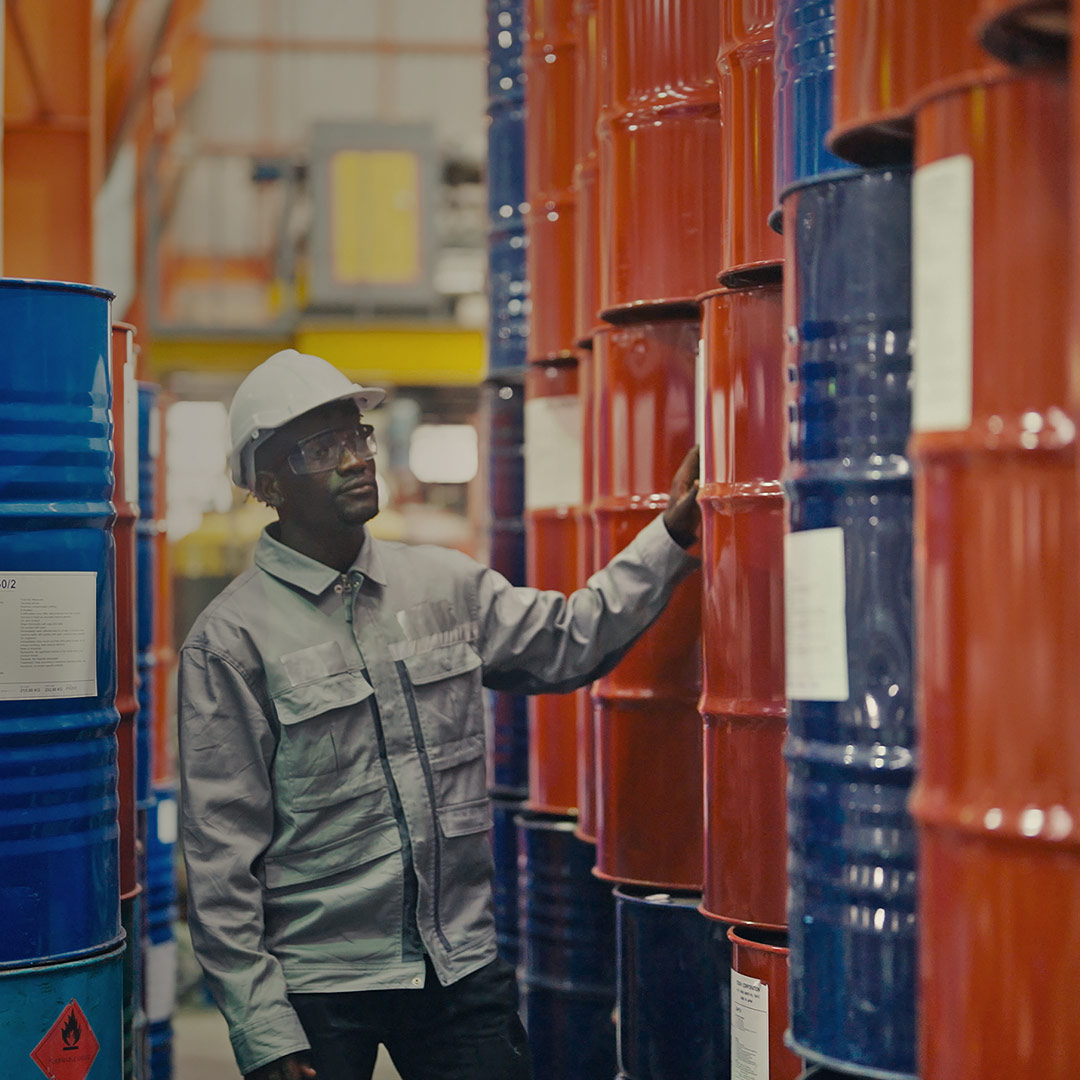 Worker in hard hat and safety glasses inspects stacked blue and red industrial barrels in a warehouse.