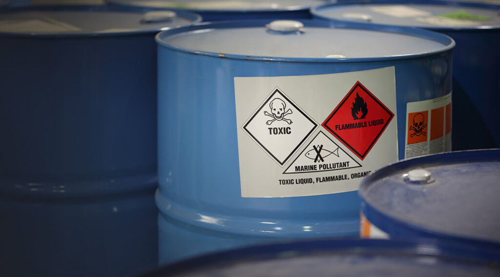 Blue metal barrels, tightly packed, with one displaying toxic, flammable liquid warnings and a marine pollutant symbol.