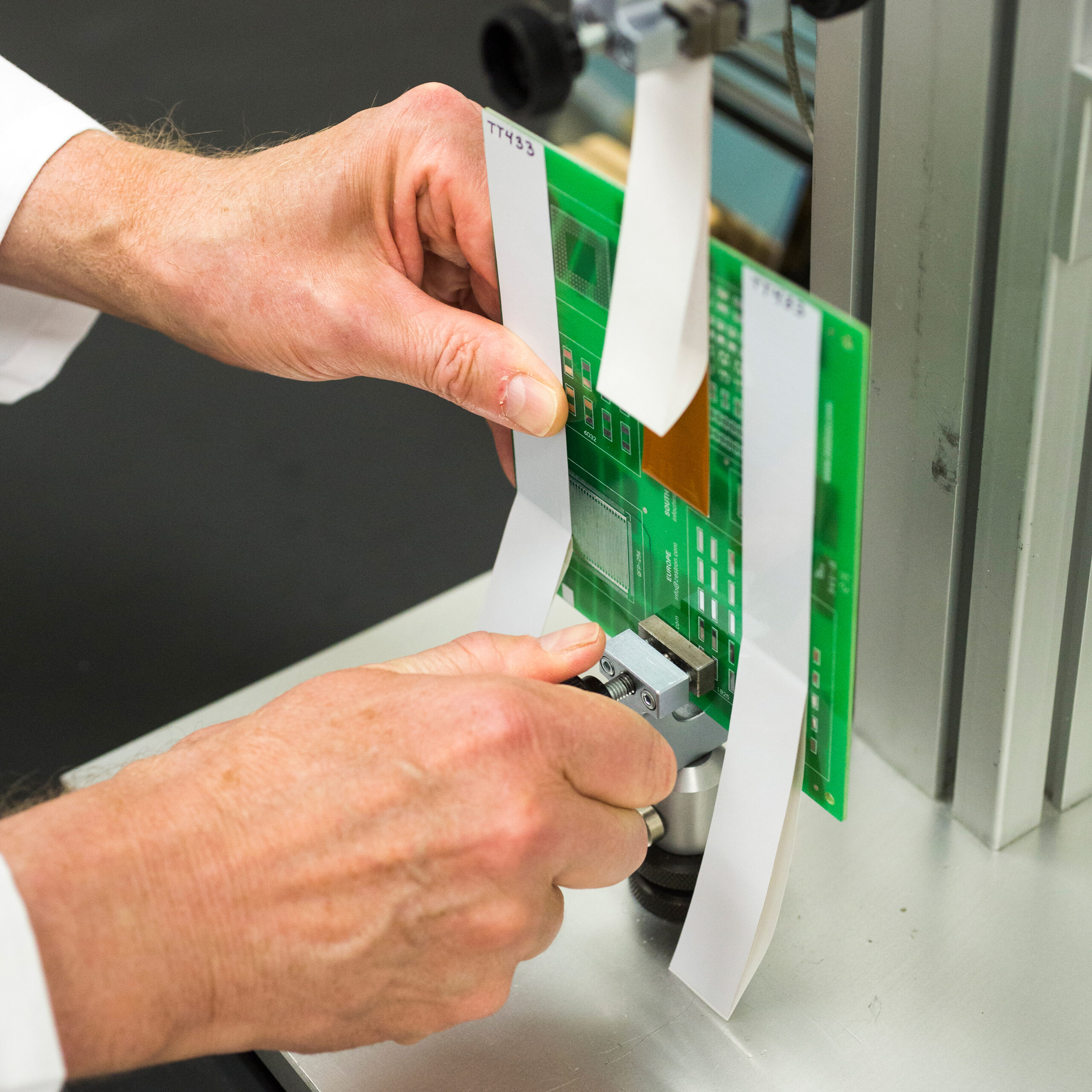 Person testing a green PCB held by two white strips on a laboratory device using a measurement tool.
