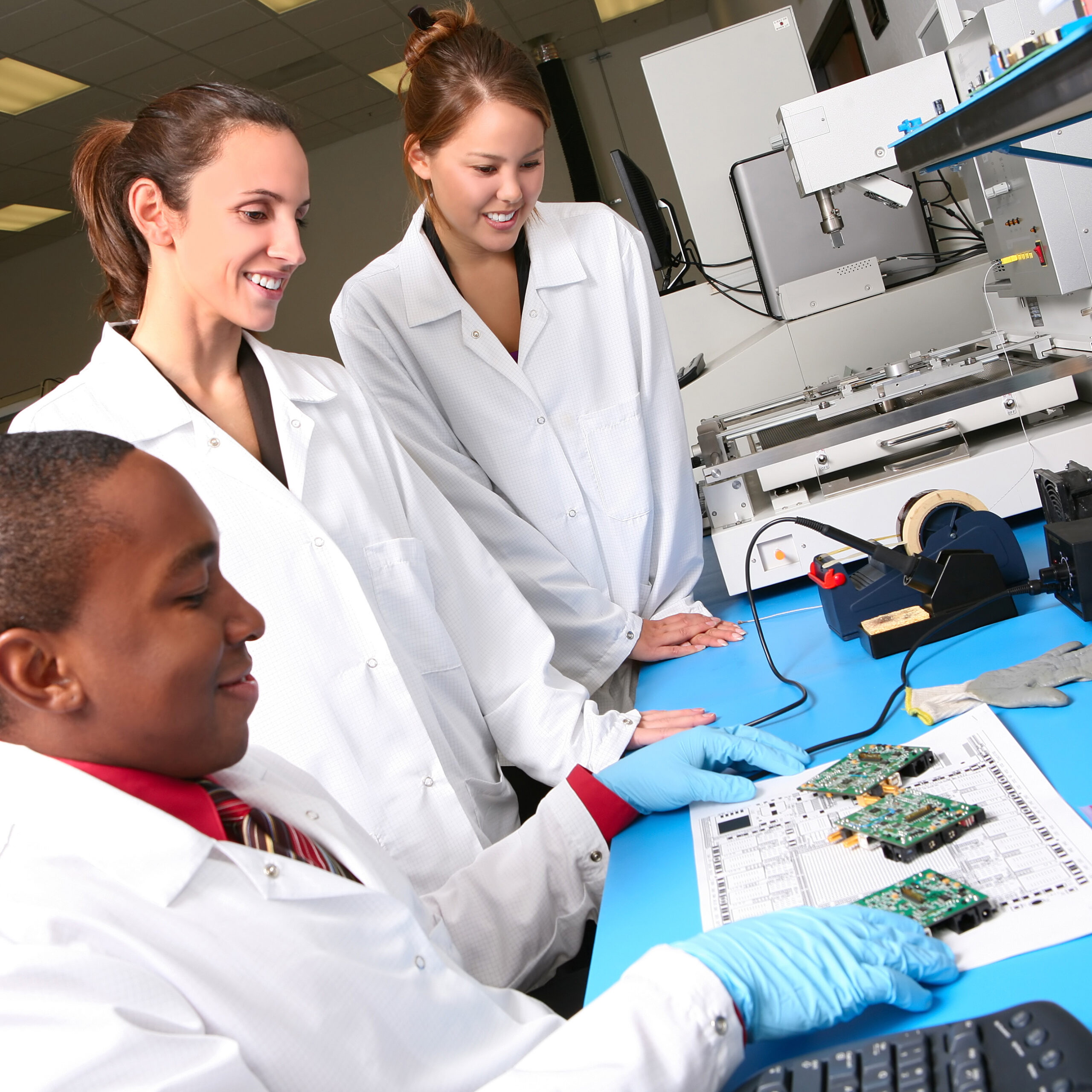 Three scientists in lab coats, one with blue gloves, study circuit boards and documents at a lab table with technical equipment nearby.