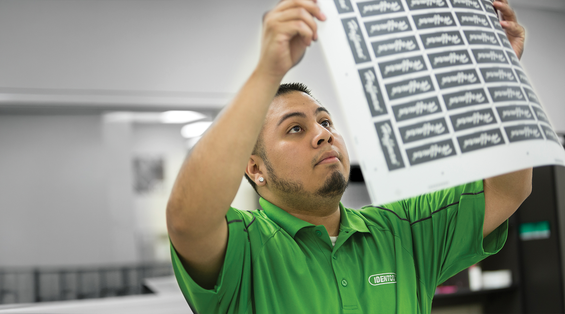 Man in green shirt examines a printed sheet with multiple labels, checking print quality closely in a print shop environment.