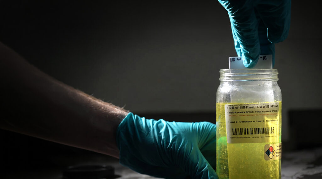 Gloved hands dip a test strip into a clear jar of yellow liquid with a warning label, barcodes, and hazard symbols for analysis.