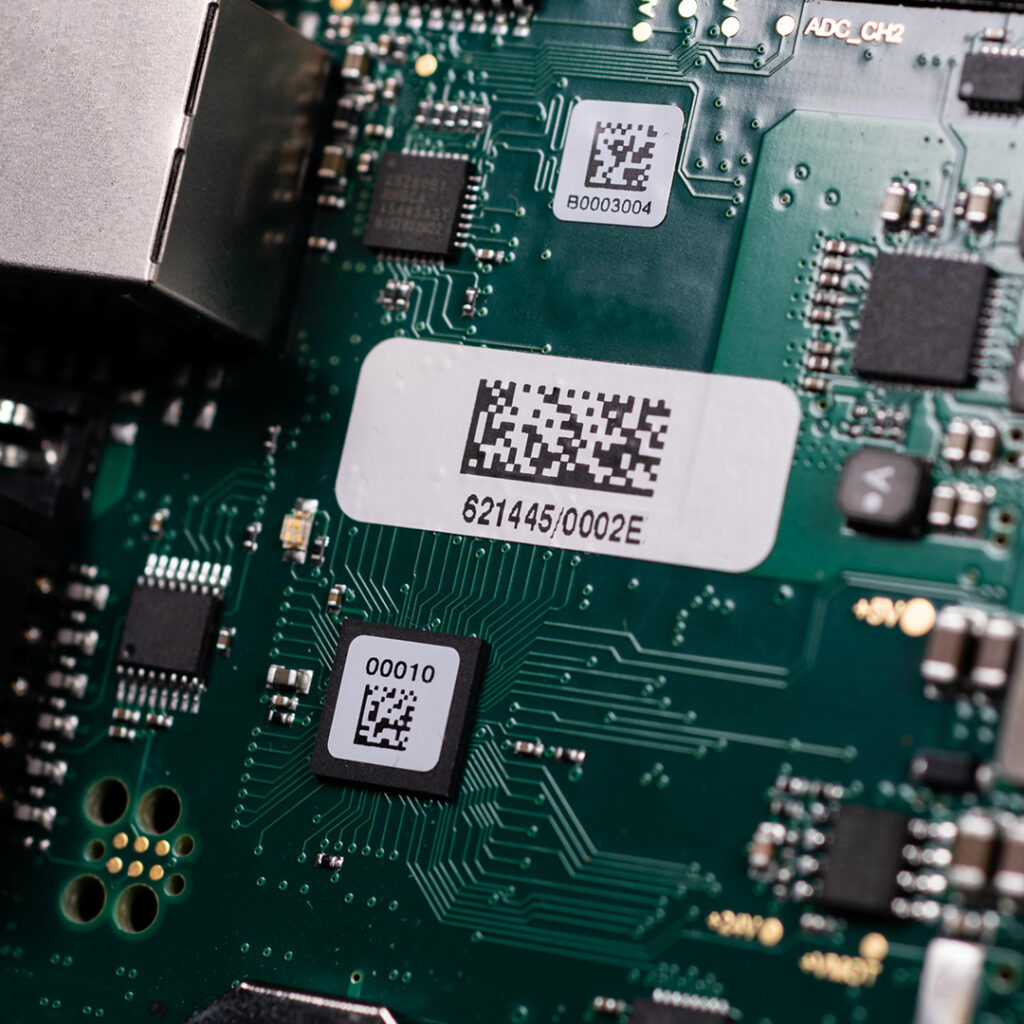 Close-up of green circuit board featuring microchips, QR codes, printed codes, electronic components, and intricate traces.