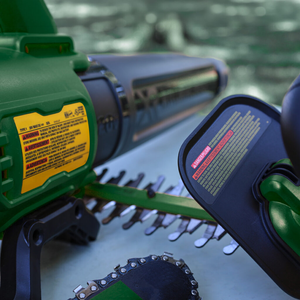 Green electric hedge trimmer with yellow and red warning labels, sharp blades visible, blurred greenery in the background.