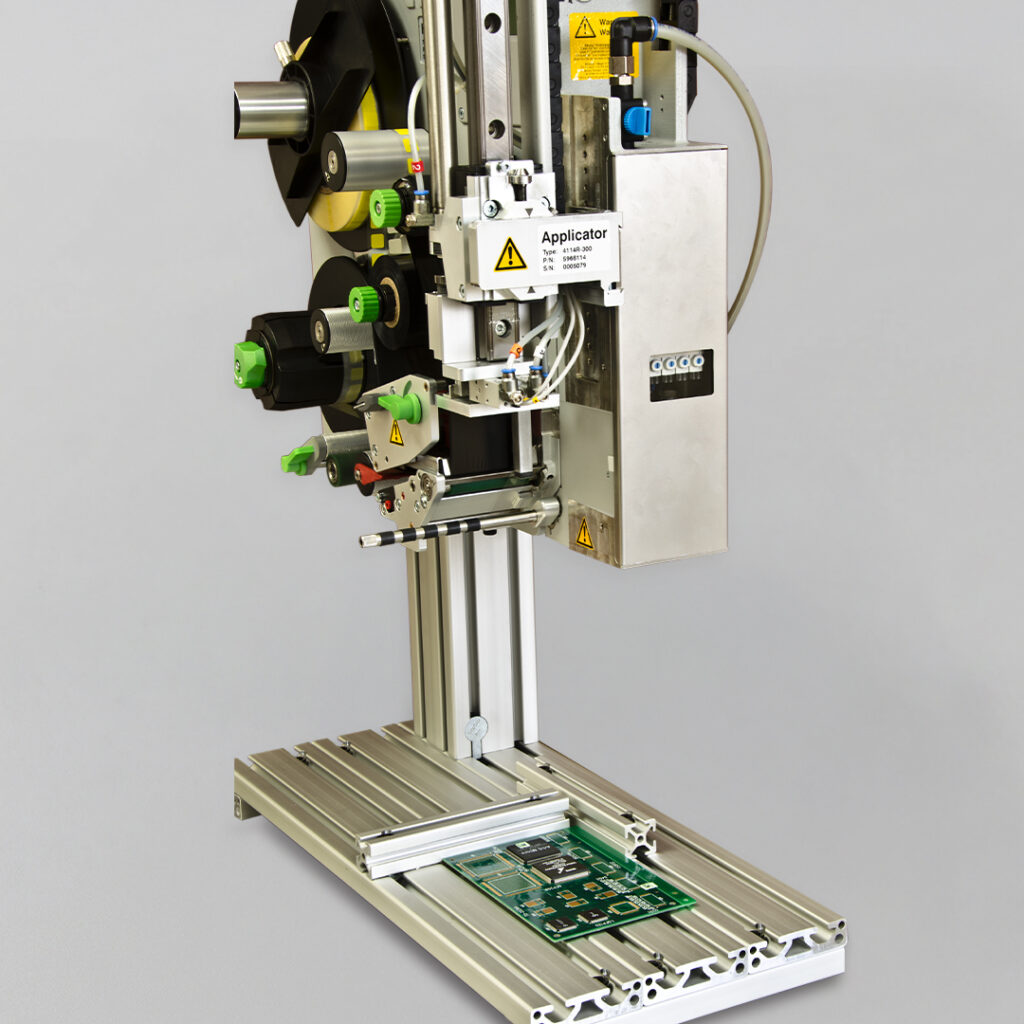 Automated labeling machine with control panel and applicator arm above green circuit board on metal base, gray background.