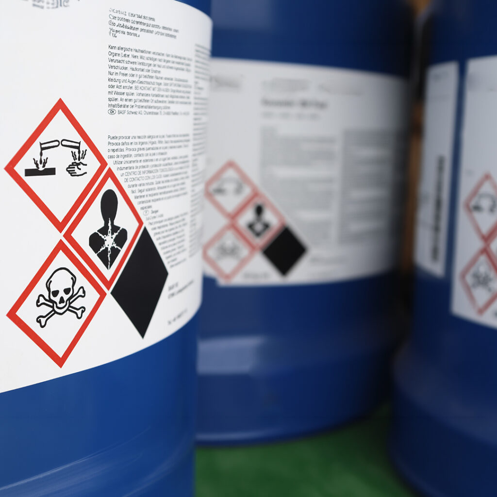 Blue chemical barrels with clear hazard labels showing corrosive, toxic, and health risk symbols and readable safety text.