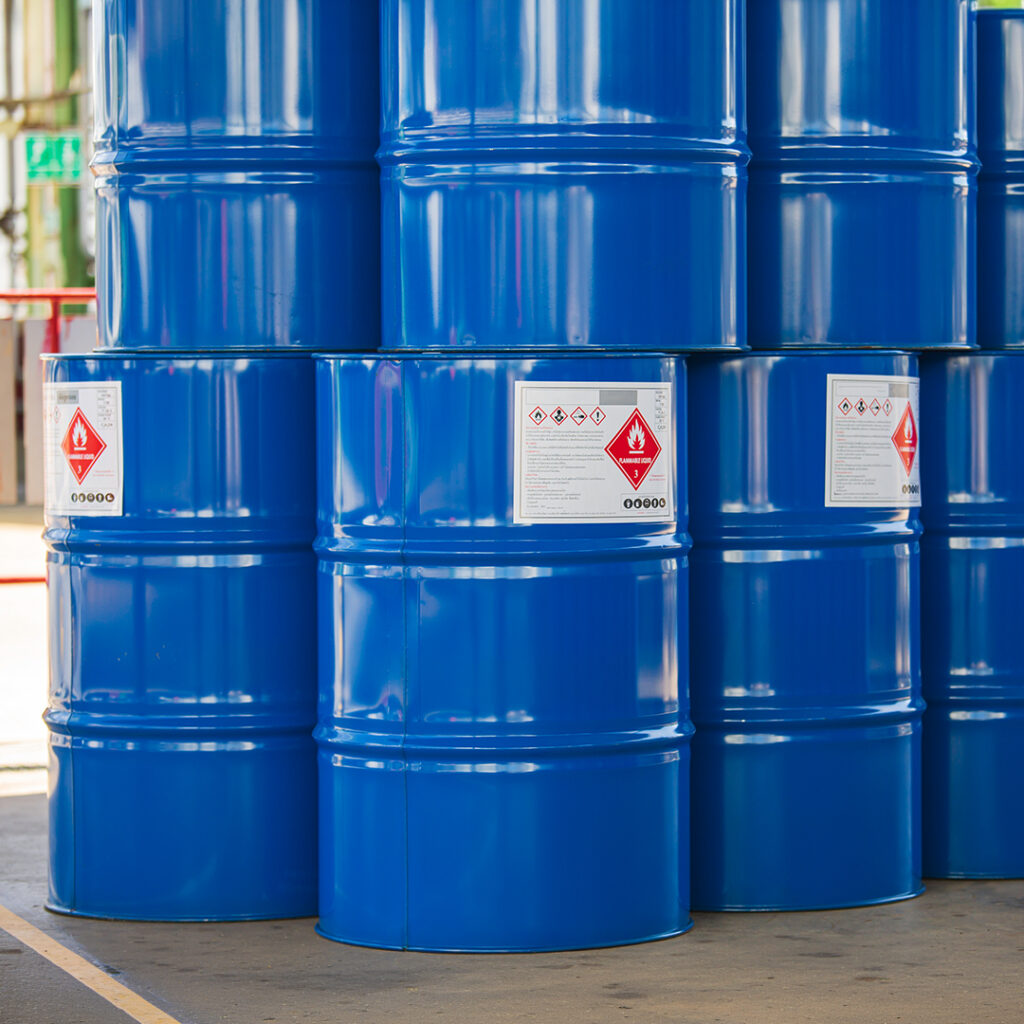 Shiny blue metal barrels with hazardous material warning labels stacked outdoors in bright sunlight.
