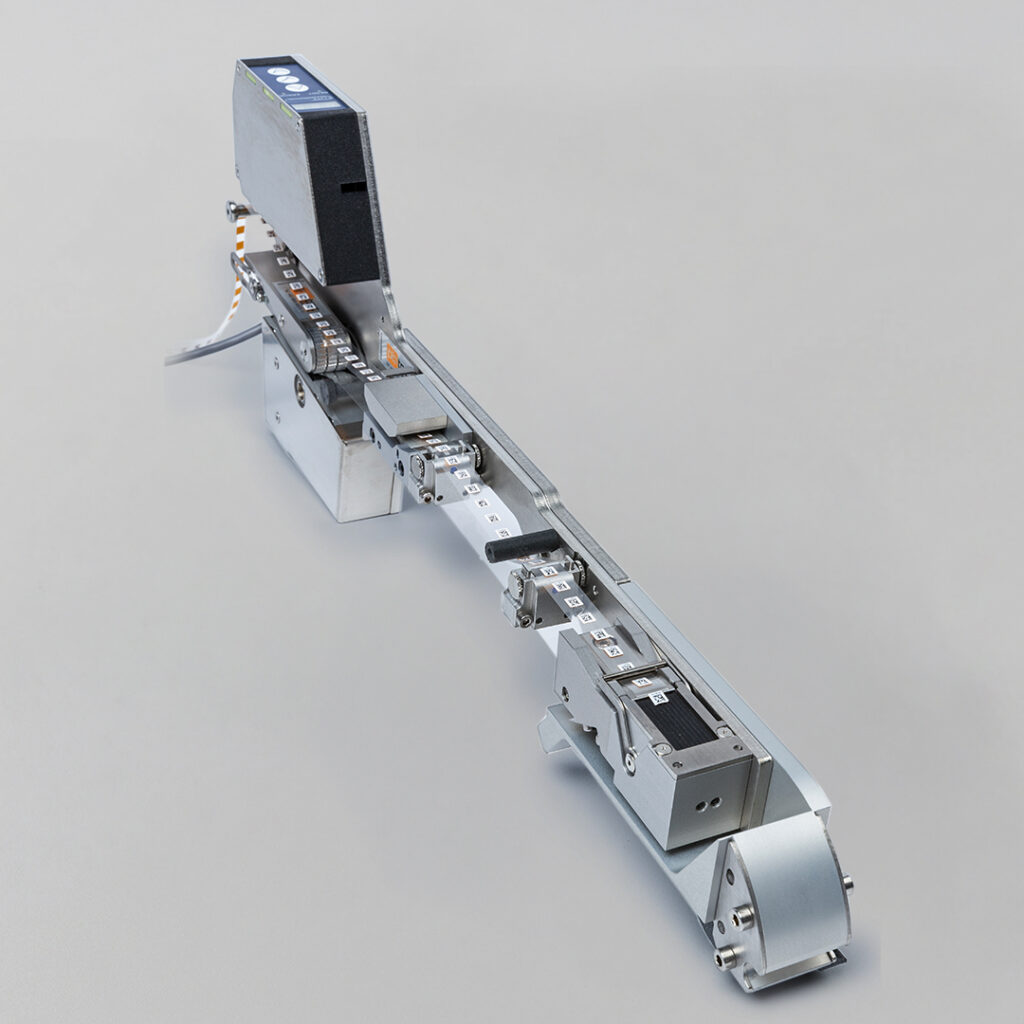 Metallic industrial machine with electronic parts, black control box, conveyor belt, labeled buttons or sensors on gray background.