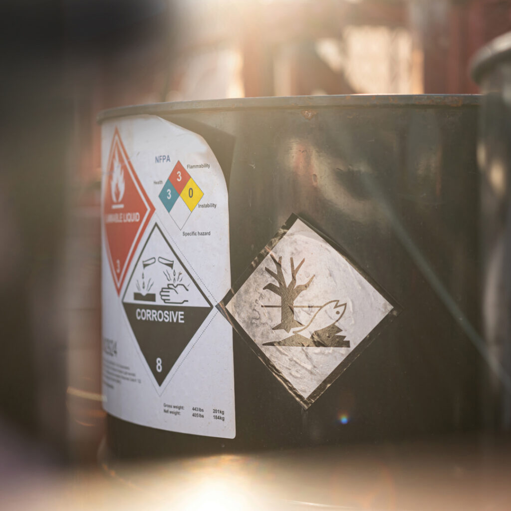 Metal drum in sunlight displaying hazard labels for flammable liquid, environmental hazard, and corrosive material.