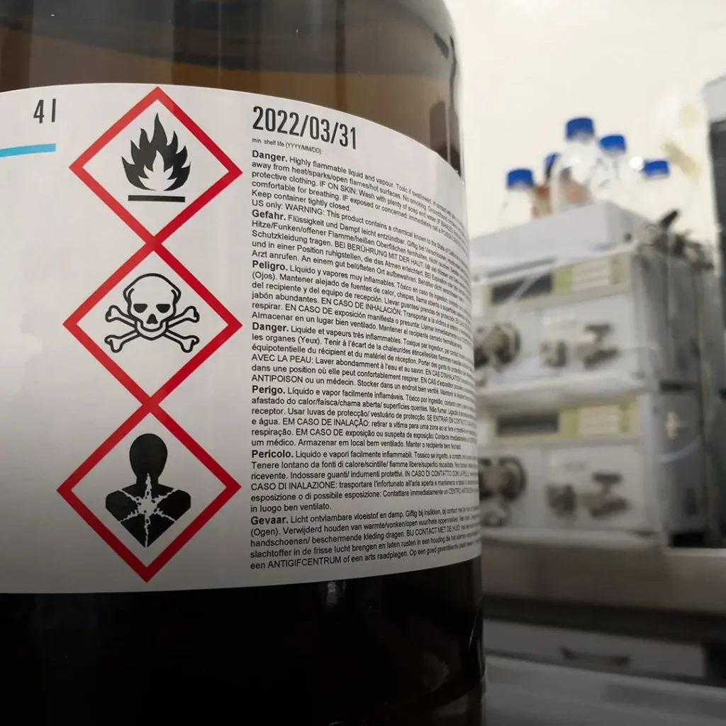 Chemical bottle label showing flammable, toxic, and health hazard symbols with blurred lab equipment in the background.