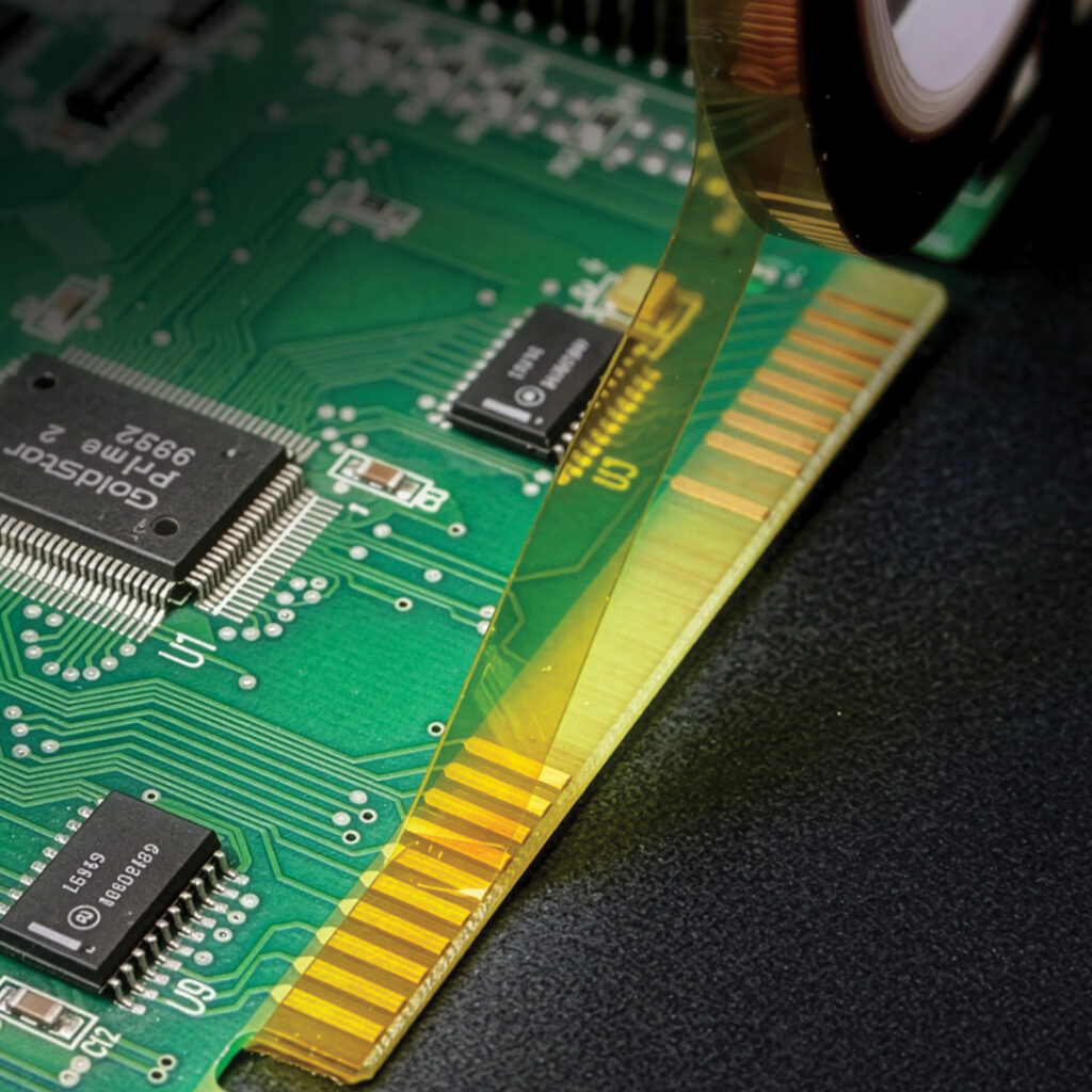 Green circuit board close-up with electronic components and yellow film peeling off gold connector pins at the edge.