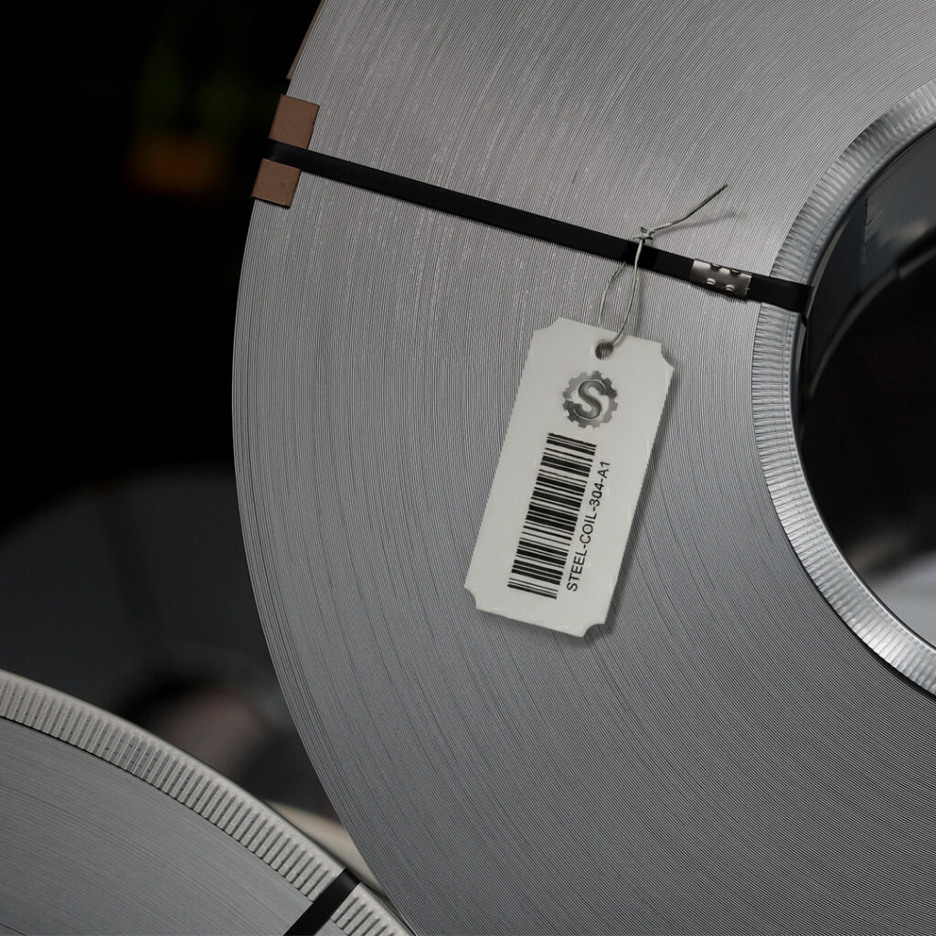 Large steel coil secured with a black strap, showing a price tag with barcode, dollar sign, and label STEEL COIL 334A41.