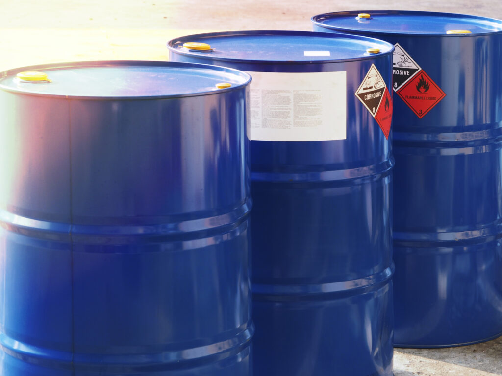 Blue metal barrels with closed lids sit on concrete outdoors, featuring corrosive and flammable hazard labels and some white tags.