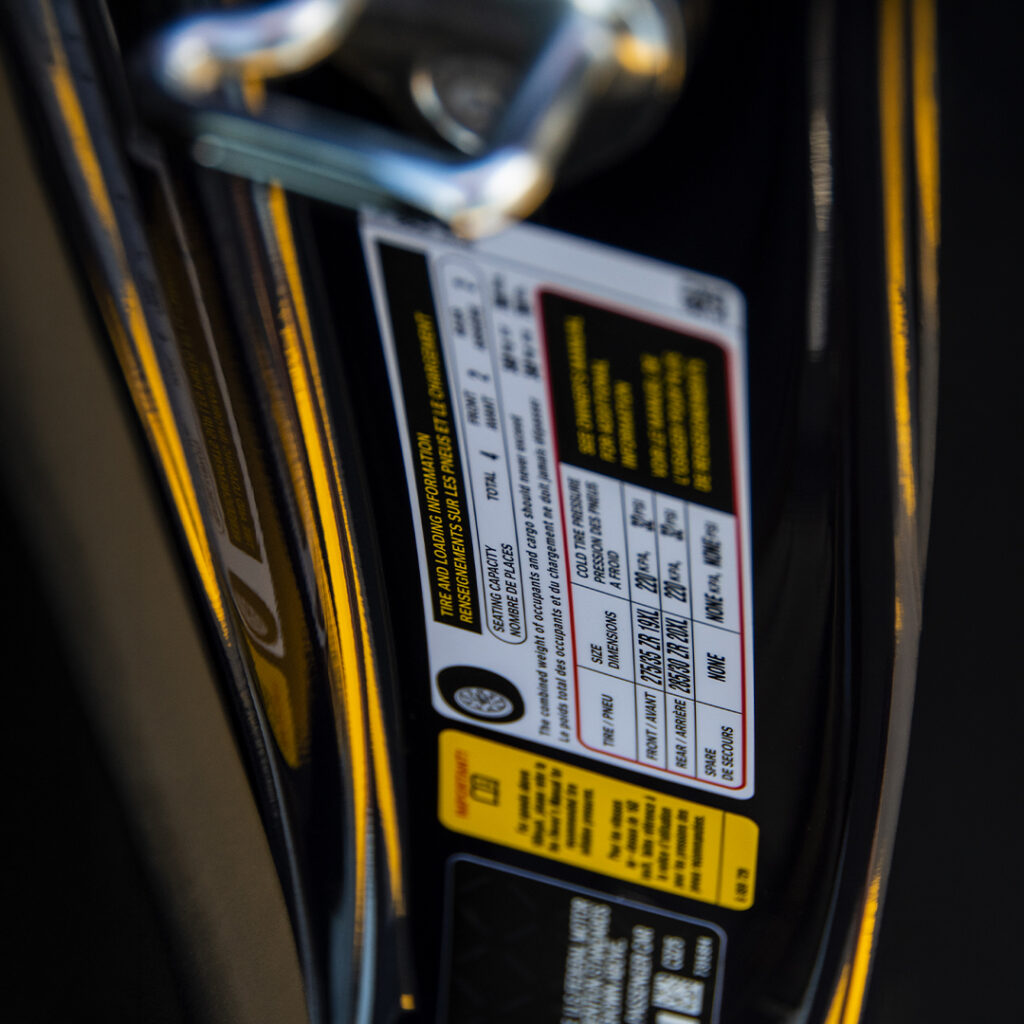 Close-up of a car door sticker displaying tire sizes, pressure recommendations, and warnings in black, white, red, and yellow text.