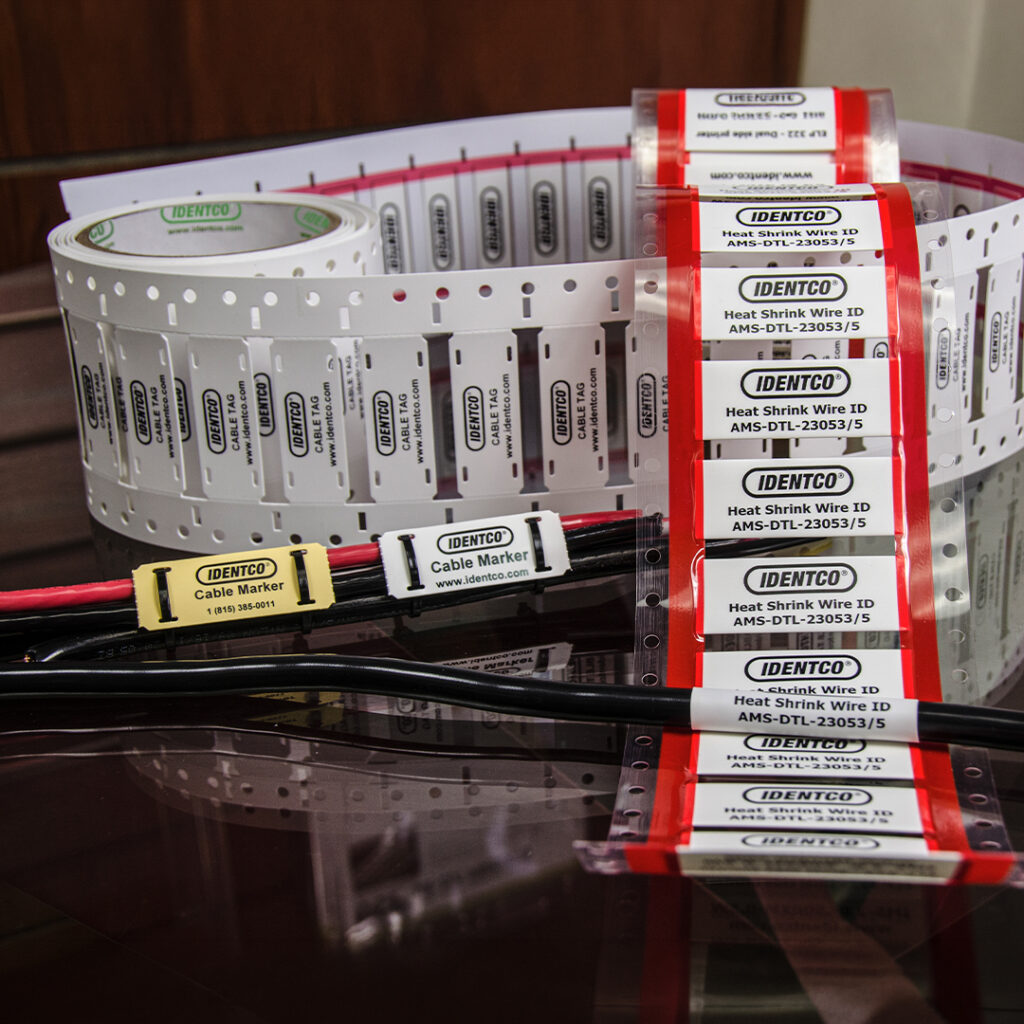 IDENTCO wire and cable markers in white, yellow, and red on rolls with sample cables fitted with markers on a reflective surface.