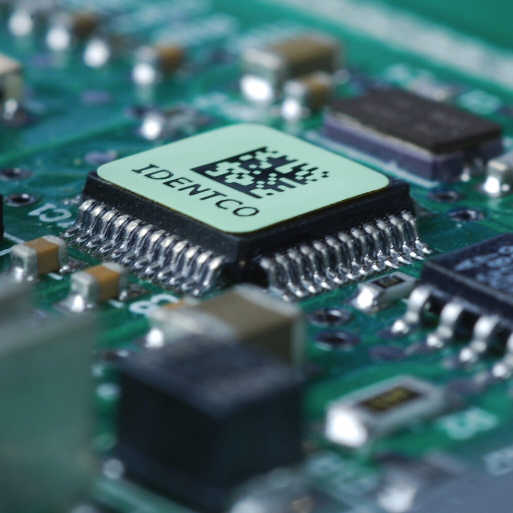Close-up of green circuit board featuring IDENTCO microchip, barcode, detailed metal pins, and surrounding electronic components.