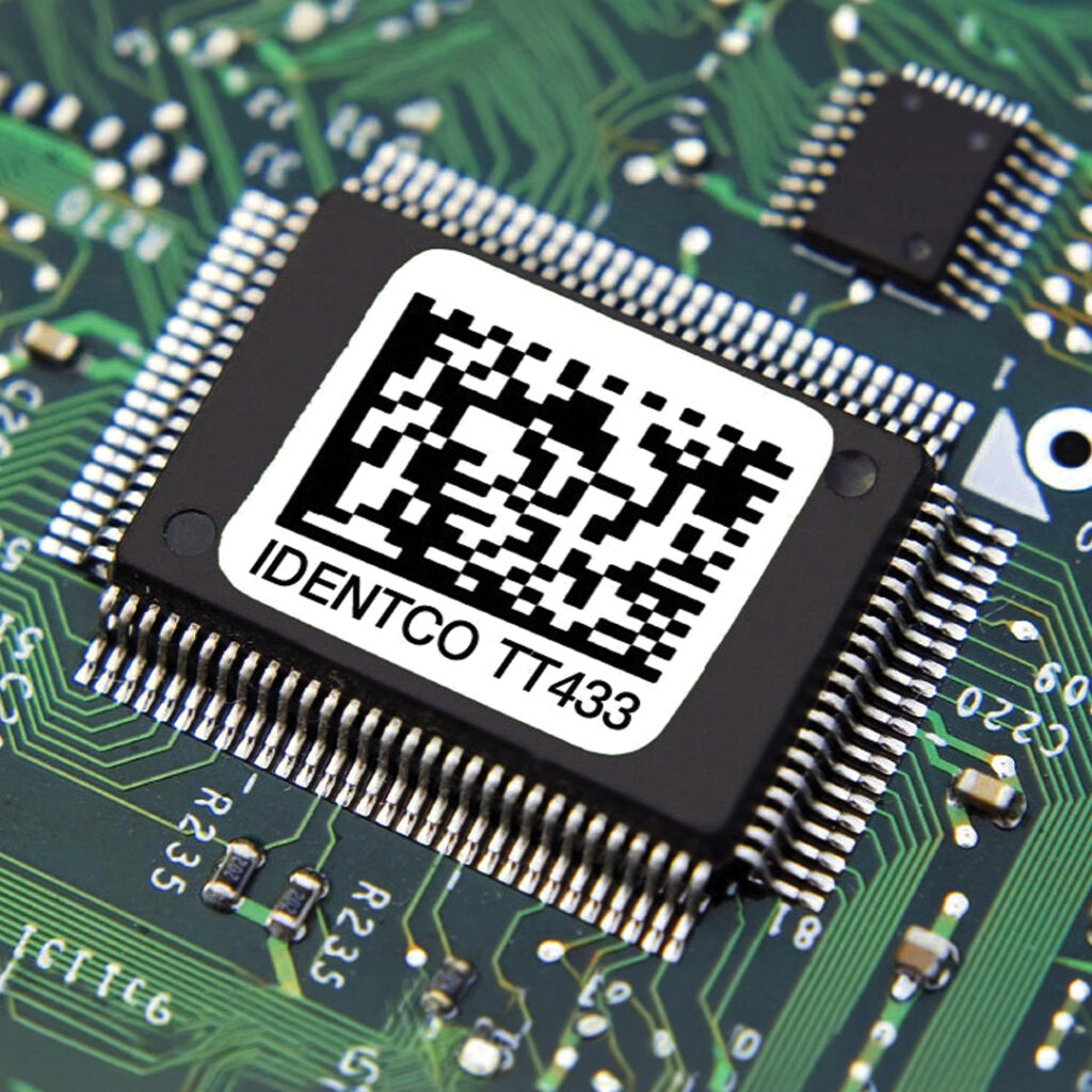Close-up of a microchip on a circuit board with a white IDENTCO T7433 label and black barcode underneath.
