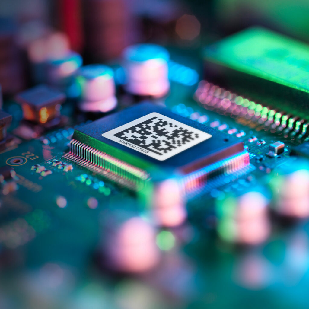 Close-up of a computer chip with a QR code on its surface, mounted on a circuit board and lit by colorful lights.