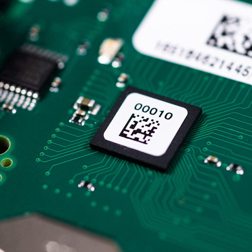 Close-up green circuit board showing a microchip with white label 00010, QR code, and surrounding electronic pathways and components.