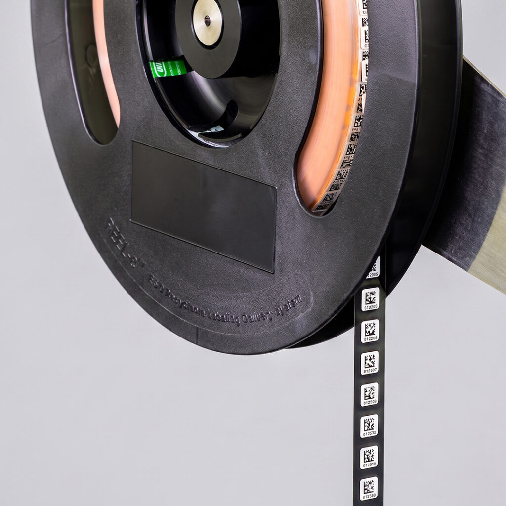 Black spool with tape reel holding small components, each marked with a QR code, on a light gray background.