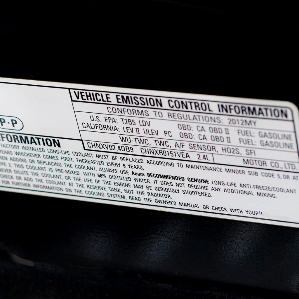 White sticker under car hood displaying vehicle emission control info, engine codes, and maintenance instructions in black text.