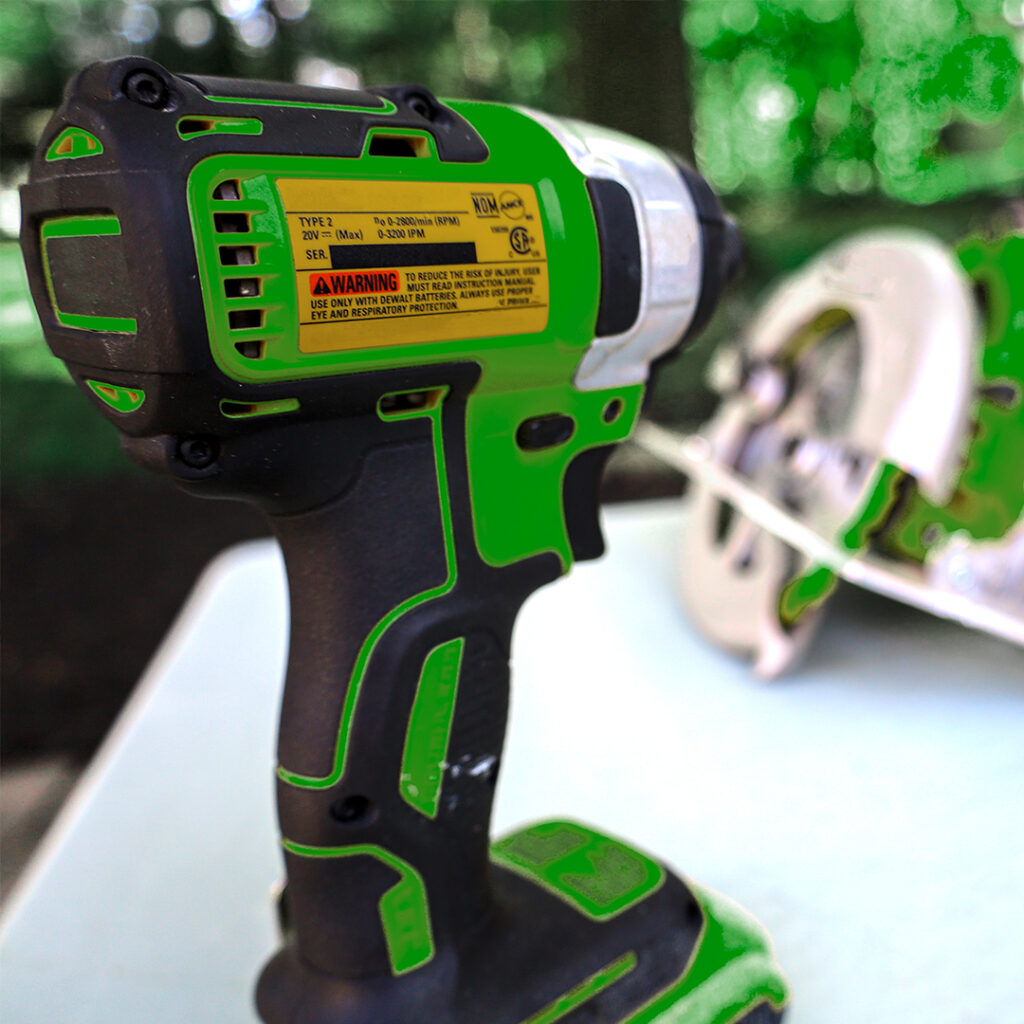 Green and black cordless power drill with yellow warning label on white outdoor surface, blurred trees and tool in background.