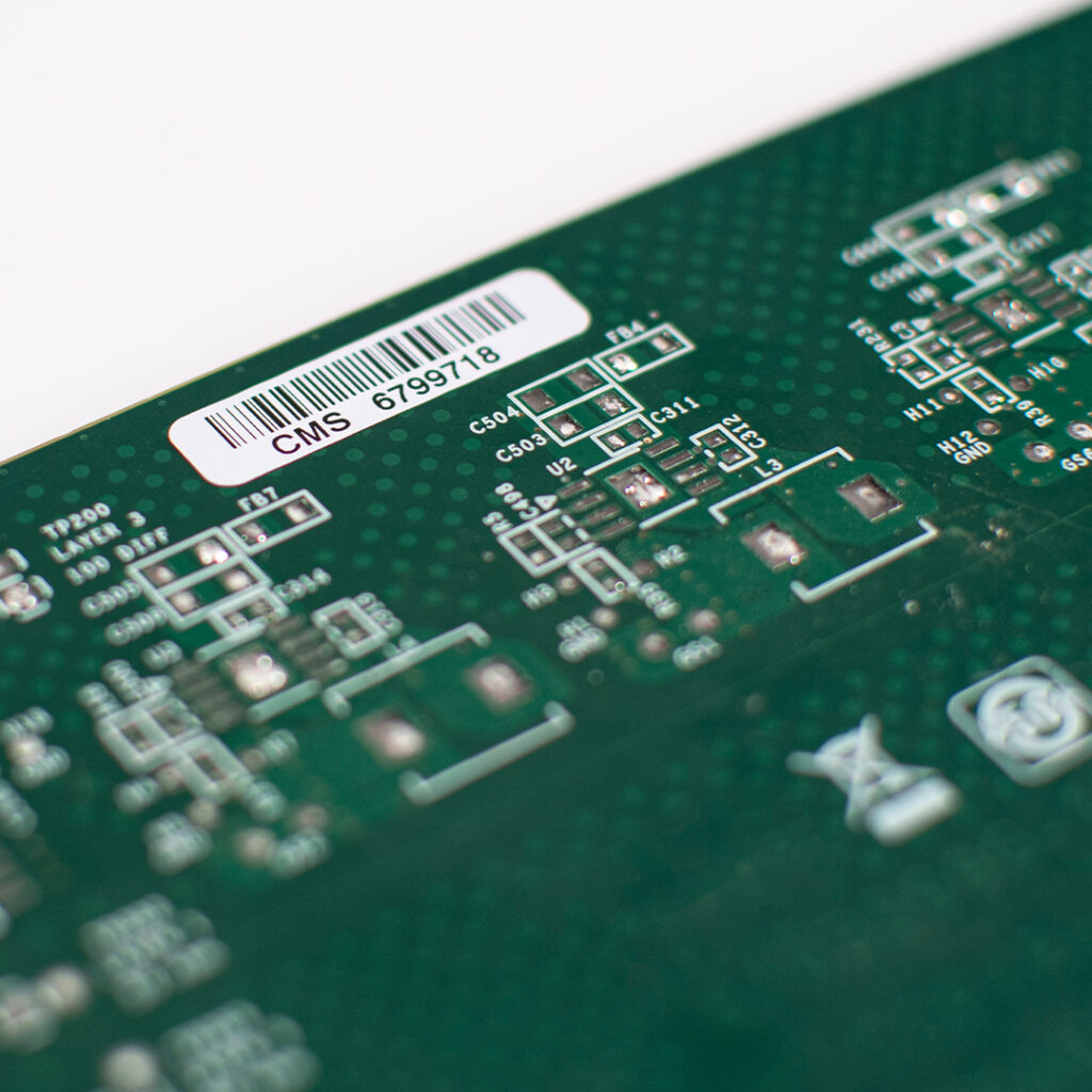 Green circuit board close-up showing white labels, electronic component outlines, circuit traces, and a barcode sticker CMS 6799718.