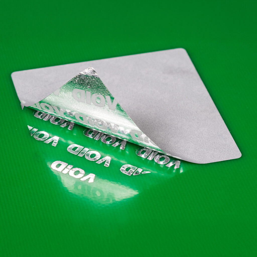 Partially peeled silver security sticker on green surface, exposing repeated VOID pattern as evidence of tampering.