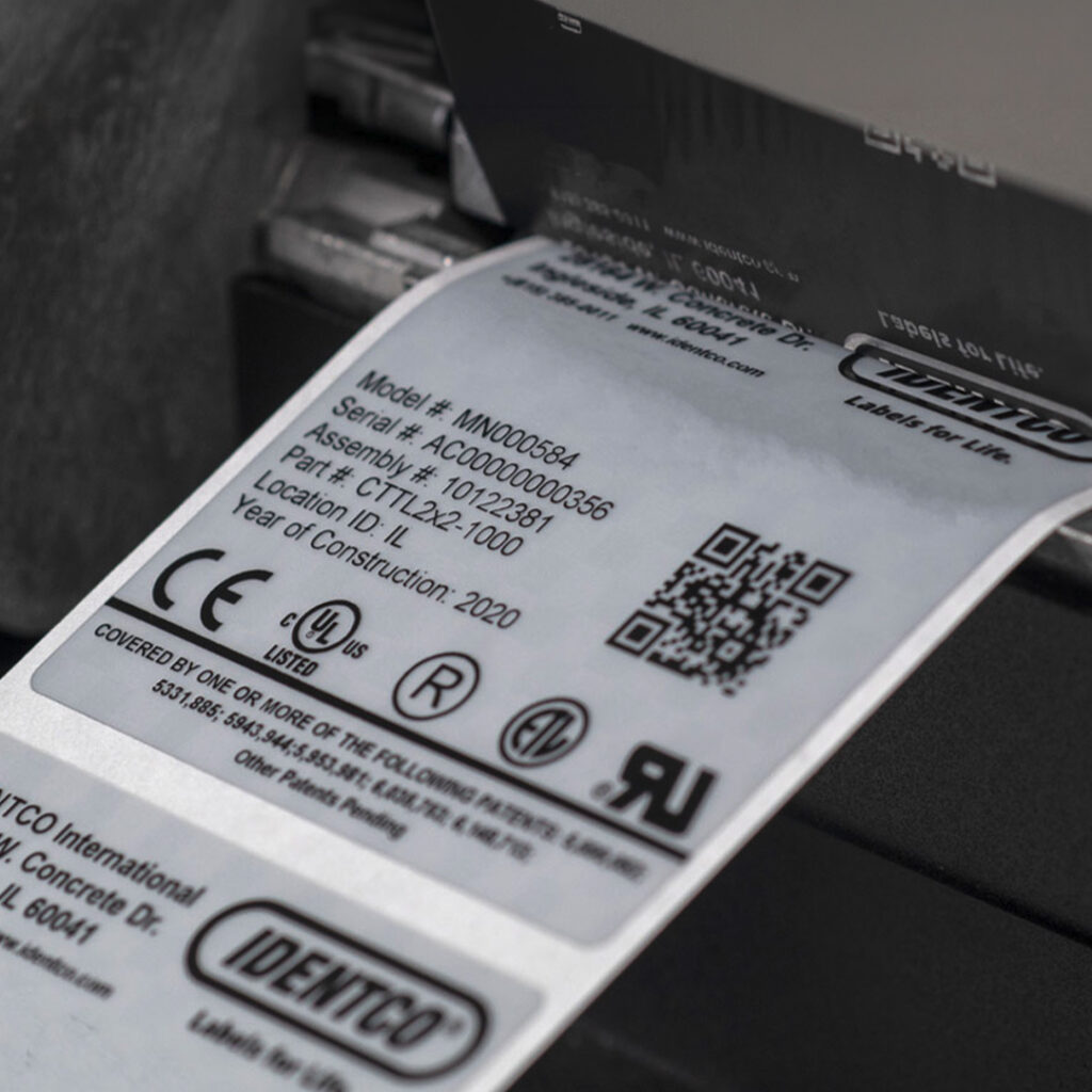 Label printer printing a white label with technical details, certifications, QR code, and Identco logo visible.